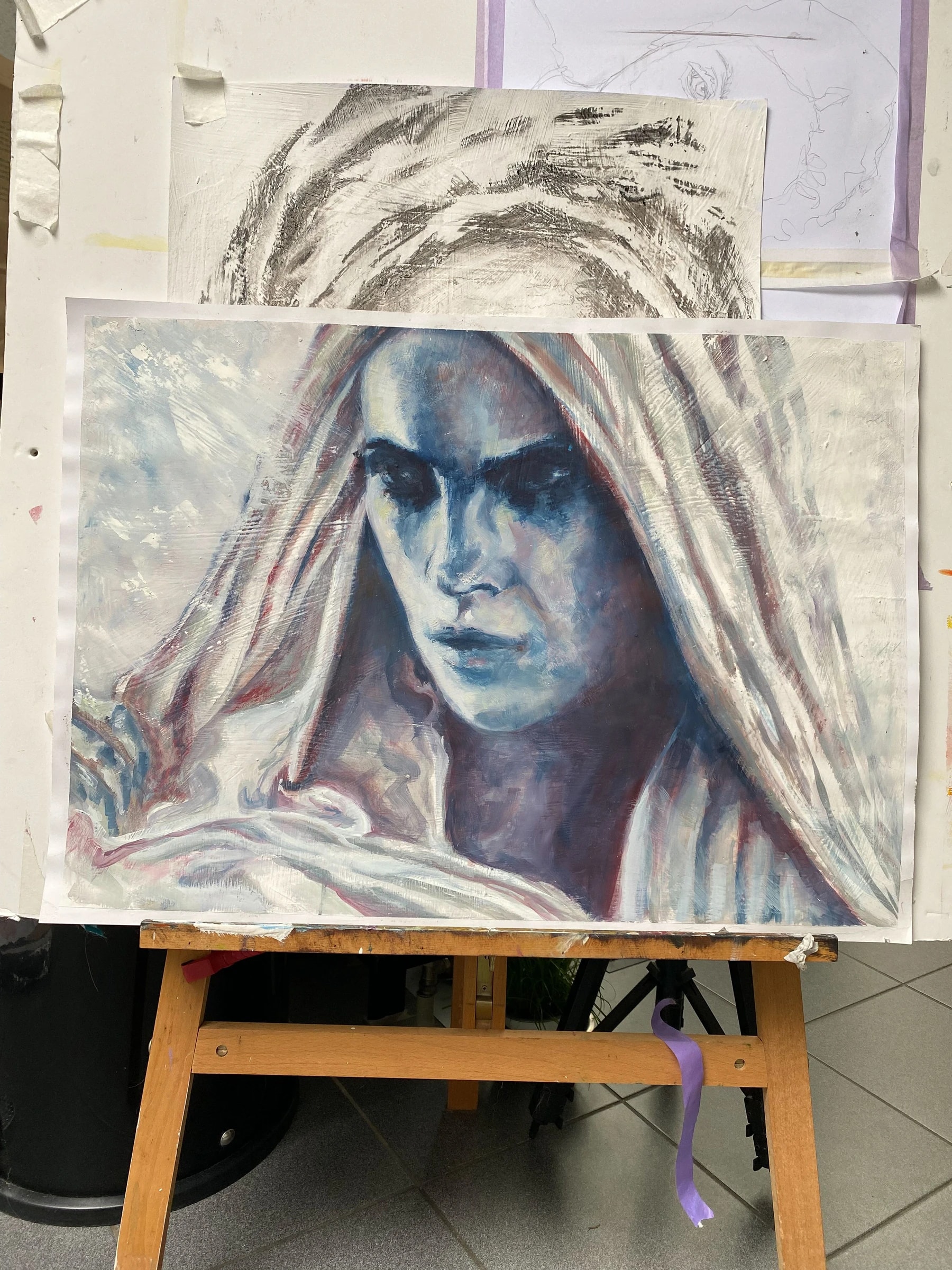 Painting on a wooden studio easel of a hooded head in cold blue and plum with dark eyes, an earlier charcoal version of the motif pinned behind it.
